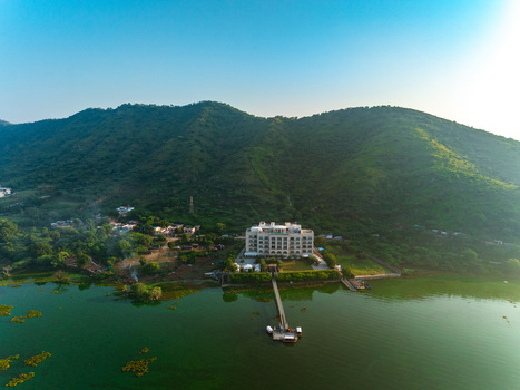 Aerial view of a luxury hotel situated on the banks of a serene lake surrounded by lush green hills under a clear sky in Udaipur Rajasthan India. Aerial view of a luxury hotel situated on the banks of a serene lake surrounded by lush green hills under a clear sky in Udaipur Rajasthan India.