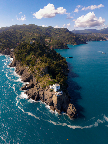 Aerial view of the Portofino lighthouse and rocky promontory surrounded by turquoise Mediterranean waters under a blue sky with white clouds in Portofino Liguria Italy.