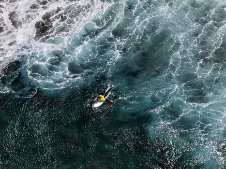 Aerial view of a surfer in a yellow wetsuit paddling through white ocean foam and dark blue waves at Praia do Areal de Santa Barbara in Ribeira Grande Acores Portugal. Aerial view of a surfer in a yellow wetsuit paddling through white ocean foam and dark blue waves at Praia do Areal de Santa Barbara in Ribeira Grande Acores Portugal.