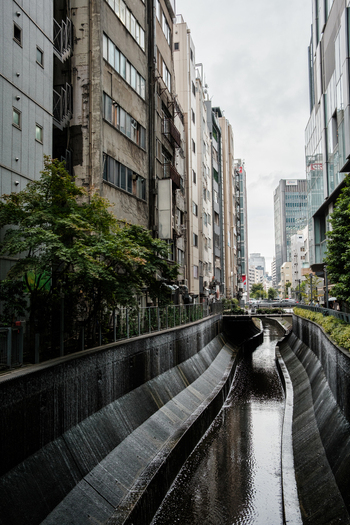 View of a narrow canal lined with concrete banks reflecting the muted sky shadowed by towering buildings in a modern urban landscape Tokyo Tokyo Japan. View of a narrow canal lined with concrete banks reflecting the muted sky shadowed by towering buildings in a modern urban landscape Tokyo Tokyo Japan.