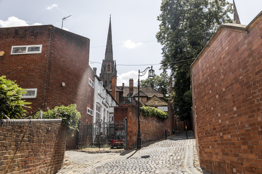 View of a narrow cobblestone street lined with historic red brick walls leading towards a church spire in Coventry England United Kingdom.