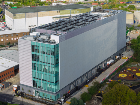 Aerial view of a modern data center building with a glass facade and rooftop cooling units situated near industrial infrastructure and a small playground in the United Kingdom. Aerial view of a modern data center building with a glass facade and rooftop cooling units situated near industrial infrastructure and a small playground in the United Kingdom.