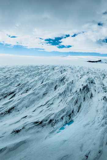 Aerial view of Vatnajokull glacier with deep crevasses textured ice formations and a small turquoise meltwater pool under a cloudy sky in Iceland.