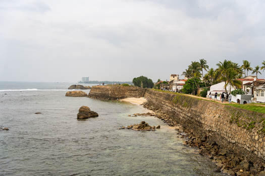 View of the historic Galle Fort ramparts with people walking along the stone walls and palm trees under an overcast sky Galle Southern Province Sri Lanka.