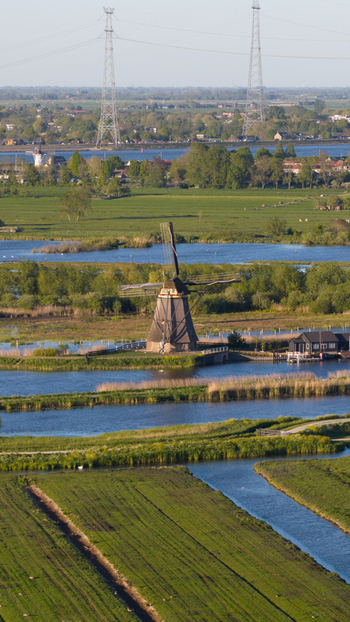 Aerial view of a historic Kinderdijk windmill standing amidst lush green fields and winding canals with power lines in the background Kinderdijk South Holland Netherlands. Aerial view of a historic Kinderdijk windmill standing amidst lush green fields and winding canals with power lines in the background Kinderdijk South Holland Netherlands.