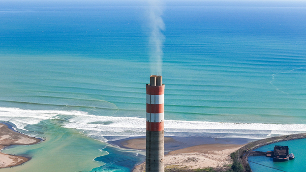 Aerial view of a tall red and white striped power plant chimney emitting smoke on the coastline with blue ocean waves and a small harbor in Indonesia.