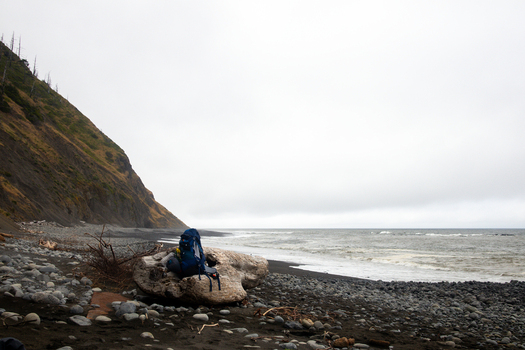 View of a blue backpack on a large piece of driftwood along the rugged shoreline of the Lost Coast with steep cliffs and crashing waves Humboldt County California United States.