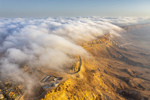 Aerial view of Mitzpe Ramon crater with a thick layer of clouds spilling over the edge of the desert cliffs illuminated by golden hour sunlight in Mitzpe Ramon South District Israel. Aerial view of Mitzpe Ramon crater with a thick layer of clouds spilling over the edge of the desert cliffs illuminated by golden hour sunlight in Mitzpe Ramon South District Israel.