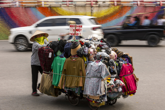 Hanoi Vietnam 11 November 2025 View of a street vendor in a conical hat pushing a cart loaded with colorful clothing along a busy street with a vibrant mural.