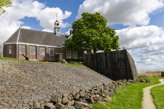 View of the historic church of Schokland with its bell tower and a large green tree behind a stone embankment under a cloudy sky in Schokland Flevoland Netherlands.