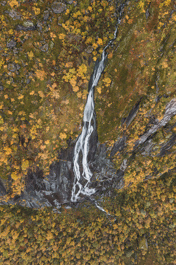 Aerial view of a waterfall cascading down a rocky cliff surrounded by autumn foliage in the Lofoten region Leknes Nordland Norway. Aerial view of a waterfall cascading down a rocky cliff surrounded by autumn foliage in the Lofoten region Leknes Nordland Norway.