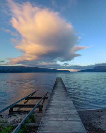 View of Lake Tarawera with a wooden jetty extending into the calm water under a dramatic sunset sky with clouds in Rotorua Bay of Plenty Region New Zealand. View of Lake Tarawera with a wooden jetty extending into the calm water under a dramatic sunset sky with clouds in Rotorua Bay of Plenty Region New Zealand.