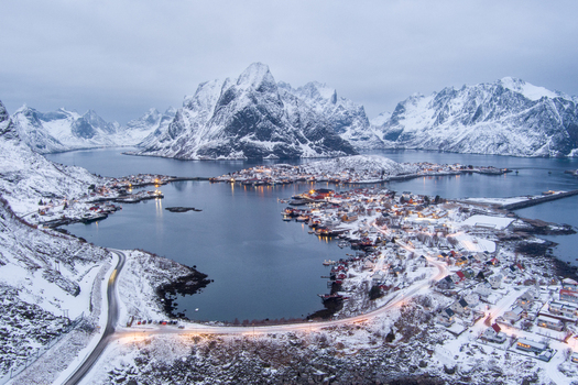 Aerial view of the snow covered village of Reine in the Lofoten archipelago featuring traditional red cabins icy waters and dramatic mountain peaks in Nordland Norway.