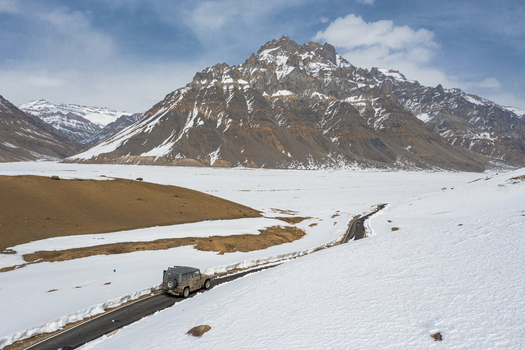 16 March 2021 Himachal Pradesh India Aerial view of a vehicle driving a mountain road in wintertime with snow capped mountain in background near Kaza township.