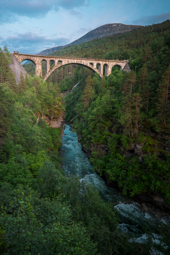 View of the historic Kylling Bridge spanning the turquoise Rauma River surrounded by lush pine forests and steep mountains in Rauma Municipality More og Romsdal Norway. View of the historic Kylling Bridge spanning the turquoise Rauma River surrounded by lush pine forests and steep mountains in Rauma Municipality More og Romsdal Norway.