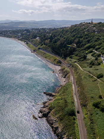 Aerial view of the Dalkey coastline featuring a train traveling along the tracks on the cliffs with lush green hills and the sea in Dublin County Dublin Ireland. Aerial view of the Dalkey coastline featuring a train traveling along the tracks on the cliffs with lush green hills and the sea in Dublin County Dublin Ireland.