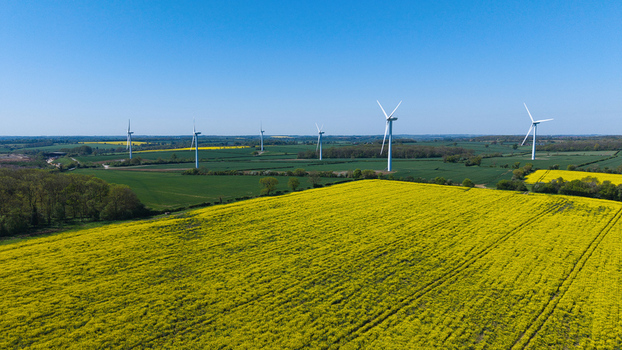 Aerial view of New Albion Wind Farm with white wind turbines standing in vibrant yellow rapeseed fields and green meadows under a clear blue sky Rushton England United Kingdom. Aerial view of New Albion Wind Farm with white wind turbines standing in vibrant yellow rapeseed fields and green meadows under a clear blue sky Rushton England United Kingdom.