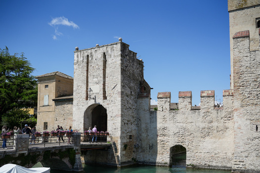 View of the historic Scaligero Castle entrance with people crossing the stone bridge over the water under a clear blue sky in Sirmione Lombardy Italy.