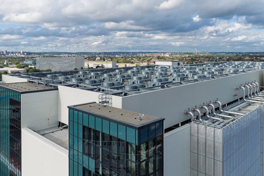Aerial view of a modern data center with numerous cooling fans on the roof and industrial piping set against an urban landscape in the United Kingdom. Aerial view of a modern data center with numerous cooling fans on the roof and industrial piping set against an urban landscape in the United Kingdom.