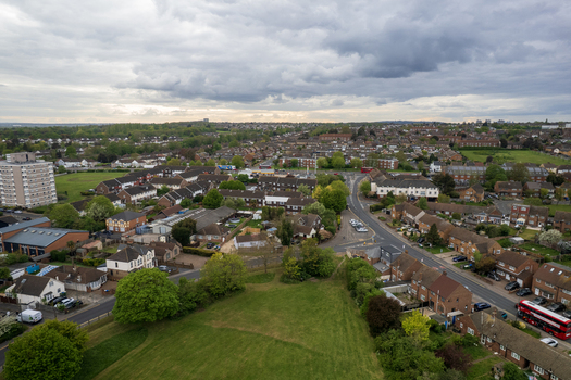 Aerial view of red double decker bus winding through streets amidst verdant trees and rooftops under a cloudy sky Slade green United Kingdom.