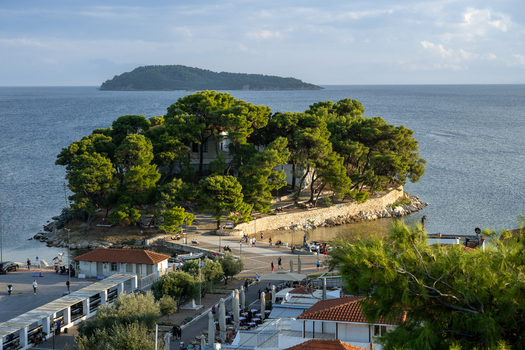 View of the island of Panagia connected by a stone bridge to the mainland with pine trees and people walking along the waterfront in Lefkada Greece.