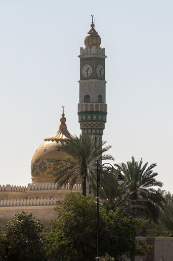 View of the ornate mosque with its golden dome and clock tower minaret rising above lush palm trees in Muscat Muscat Governorate Oman. View of the ornate mosque with its golden dome and clock tower minaret rising above lush palm trees in Muscat Muscat Governorate Oman.
