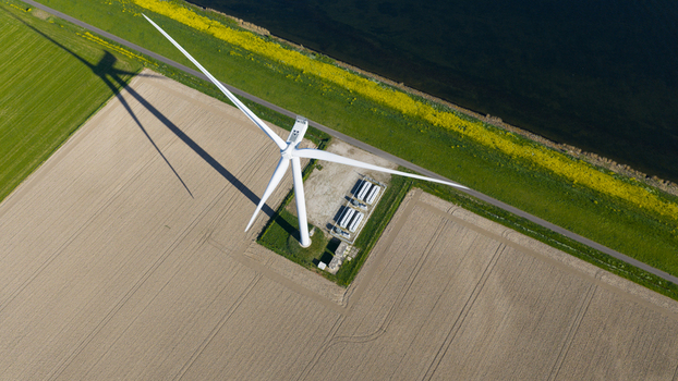 Aerial view of a large white wind turbine casting a long shadow over agricultural fields and a green dyke by the water in Ooltgensplaat Zuid Holland Netherlands. Aerial view of a large white wind turbine casting a long shadow over agricultural fields and a green dyke by the water in Ooltgensplaat Zuid Holland Netherlands.