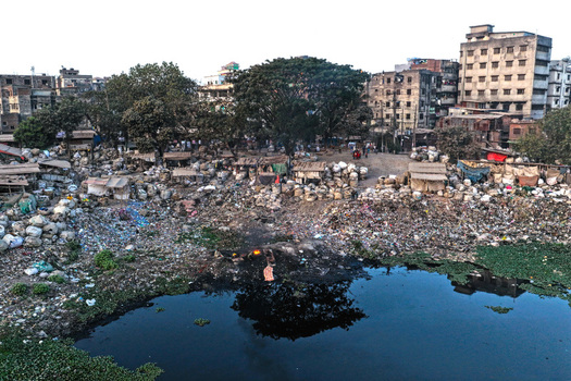 Aerial view of the polluted banks of the Buriganga River with heaps of waste makeshift shelters and urban buildings in the background in Dhaka Dhaka Division Bangladesh. Aerial view of the polluted banks of the Buriganga River with heaps of waste makeshift shelters and urban buildings in the background in Dhaka Dhaka Division Bangladesh.