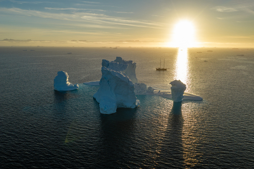 Aerial view of an arch iceberg floating in the ocean with a sailboat nearby during golden hour in Greenland. Aerial view of an arch iceberg floating in the ocean with a sailboat nearby during golden hour in Greenland.