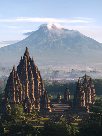 Aerial view of the ancient Prambanan Temple complex with the smoking Mount Merapi volcano in the background under a clear sky in Kecamatan Prambanan Daerah Istimewa Yogya Indonesia.