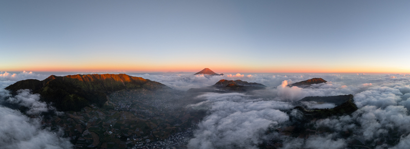 Aerial view of Dieng Plateau at sunset with volcanic peaks rising above a dense sea of clouds and terraced agricultural fields in the valley below Kejajar Central Java Indonesia.
