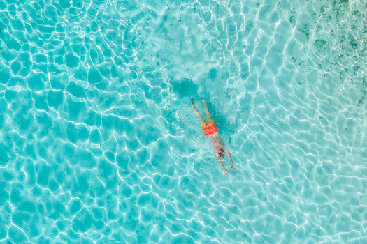 Aerial view of a man in orange shorts swimming in crystal clear turquoise water with sun ripples on the surface Innafinolhu Haa Alif Atoll Maldives. Aerial view of a man in orange shorts swimming in crystal clear turquoise water with sun ripples on the surface Innafinolhu Haa Alif Atoll Maldives.
