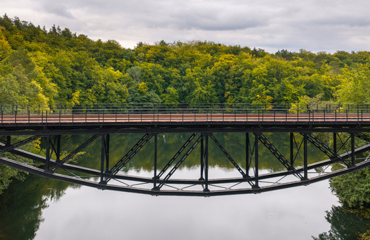 Aerial view of the historic railway truss bridge in Rutki spanning over a calm river surrounded by lush green forest in Rutki Pomeranian Voivodeship Poland. Aerial view of the historic railway truss bridge in Rutki spanning over a calm river surrounded by lush green forest in Rutki Pomeranian Voivodeship Poland.