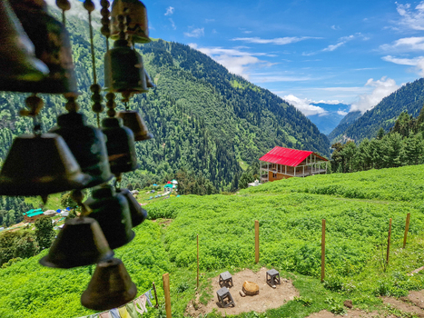 View of traditional bells in the foreground overlooking a lush green valley with a house featuring a red roof nestled in the mountains of Malana Himachal Pradesh India.