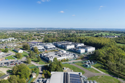 Aerial view of a large data center facility with multiple server buildings cooling units on rooftops surrounding green trees and parking areas in the United Kingdom.