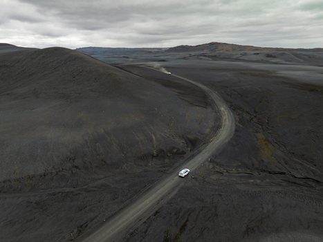 Aerial view of a white SUV driving along a winding road through a vast volcanic landscape with dark terrain under an overcast sky in Iceland. Aerial view of a white SUV driving along a winding road through a vast volcanic landscape with dark terrain under an overcast sky in Iceland.