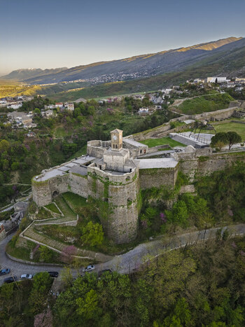 Aerial view of Gjirokaster Fortress with its iconic clock tower stone walls and surrounding lush green hills in Gjirokaster Gjirokaster County Albania. Aerial view of Gjirokaster Fortress with its iconic clock tower stone walls and surrounding lush green hills in Gjirokaster Gjirokaster County Albania.