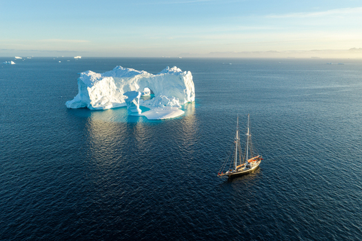Aerial view of a majestic arch iceberg floating in the deep blue Arctic waters near a classic sailing ship with calm sea and clear sky in Greenland. Aerial view of a majestic arch iceberg floating in the deep blue Arctic waters near a classic sailing ship with calm sea and clear sky in Greenland.