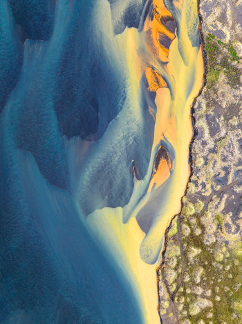 Aerial view of the vibrant glacial river delta featuring striking blue and yellow sediment patterns flowing through dark volcanic sand and mossy terrain in Iceland. Aerial view of the vibrant glacial river delta featuring striking blue and yellow sediment patterns flowing through dark volcanic sand and mossy terrain in Iceland.