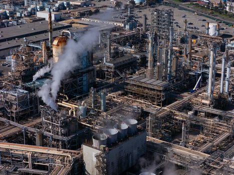 Aerial view of an industrial oil refinery in Carson featuring complex piping tall distillation towers cooling fans and steam rising from processing units in Carson California United States.