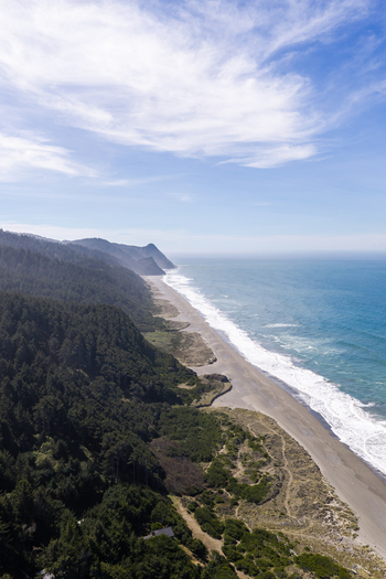 Aerial view of the rugged coastline featuring a sandy beach dense evergreen forests and crashing waves along the Pacific Ocean in Gold Beach Oregon United States. Aerial view of the rugged coastline featuring a sandy beach dense evergreen forests and crashing waves along the Pacific Ocean in Gold Beach Oregon United States.