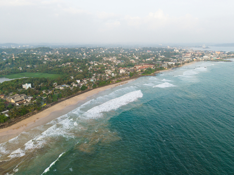 Aerial view of the Indian Ocean coastline with waves crashing on the sandy beach and urban buildings surrounded by lush greenery in Galle Southern Province Sri Lanka.
