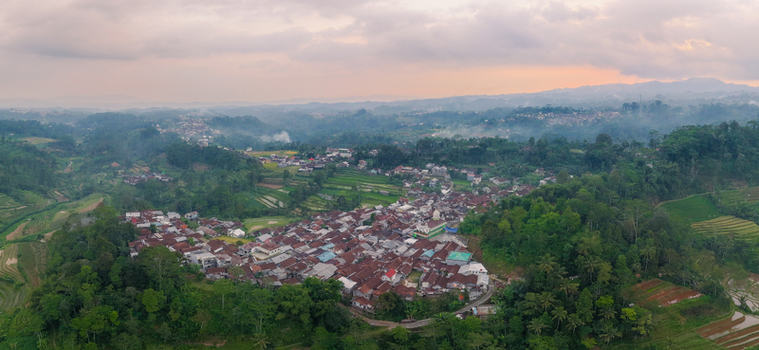 Aerial view of a rural village surrounded by lush green hills and terraced rice fields under a hazy sunset sky in Kecamatan Mojotengah Jawa Tengah Indonesia.