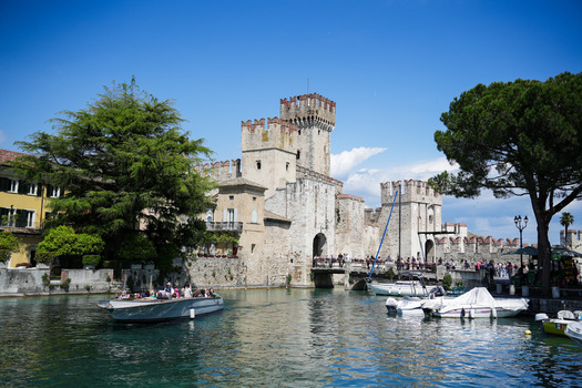 View of the medieval Scaligero Castle with its stone towers and bridge over the turquoise water of Lake Garda with tourists and boats Sirmione Lombardy Italy.
