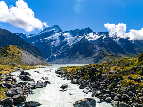 View of a glacier fed turquoise river winding its way through a rugged landscape towards snow capped mountains under a blue sky Mount Cook Canterbury Region New Zealand. View of a glacier fed turquoise river winding its way through a rugged landscape towards snow capped mountains under a blue sky Mount Cook Canterbury Region New Zealand.