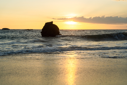 View of a large rock formation in the ocean during a golden sunset with waves crashing on the sandy beach in Galle Southern Province Sri Lanka.