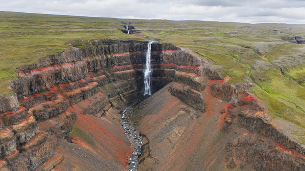 Aerial view of a cascading waterfall plunging dramatically through layered cliffs of dark basalt rock and vibrant red clay under a cloudy sky Hengifoss Iceland.