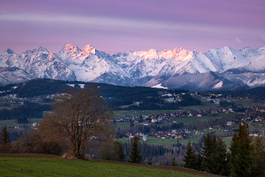 View of the snow capped Tatra Mountains rising above the village houses and green fields under a purple sunset sky in Czarna Gora Lesser Poland Poland. View of the snow capped Tatra Mountains rising above the village houses and green fields under a purple sunset sky in Czarna Gora Lesser Poland Poland.