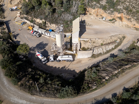 Aerial view of a concrete batching plant with cement mixers and trucks parked on a dirt site surrounded by rocky terrain and trees in Teulada Valencian Community Spain.