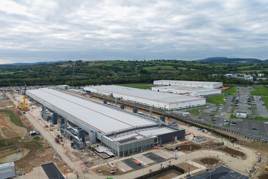 Aerial view of a large data center facility with long rectangular buildings construction equipment and parking lots surrounded by green hills in the United Kingdom.